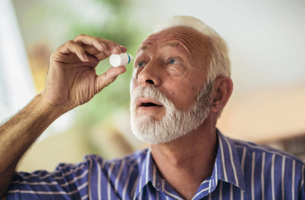 A senior male in a blue and white striped polo shirt with dry eyes is pouring some drops of eye solution into his right eye.