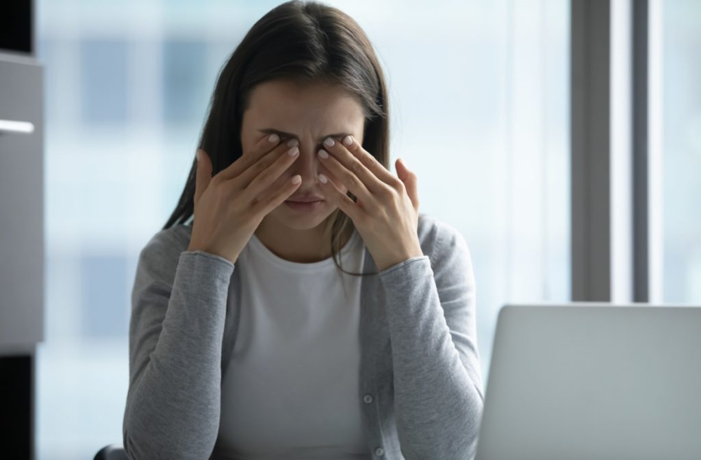 A woman sitting at a desk while working on a laptop rubs her eyes due to discomfort from increased eye pressure.