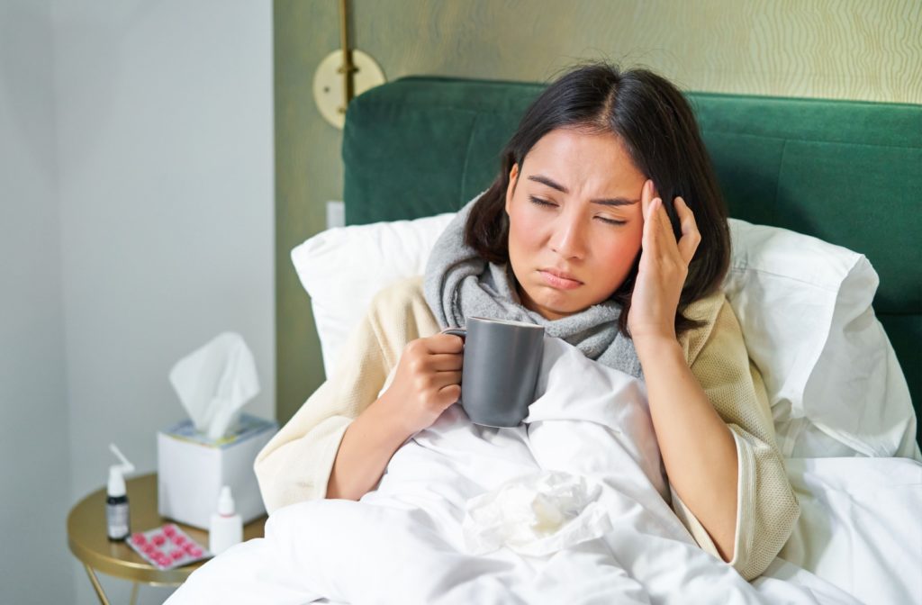 A sick person in bed with eyes closed holding a mug in one hand and placing the other on the side of their temple.