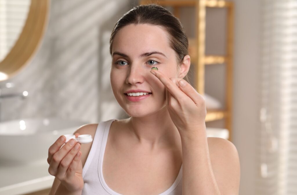 A person smiling stands in a bathroom, holding a contact lens case in one hand while applying a contact lens with the other.